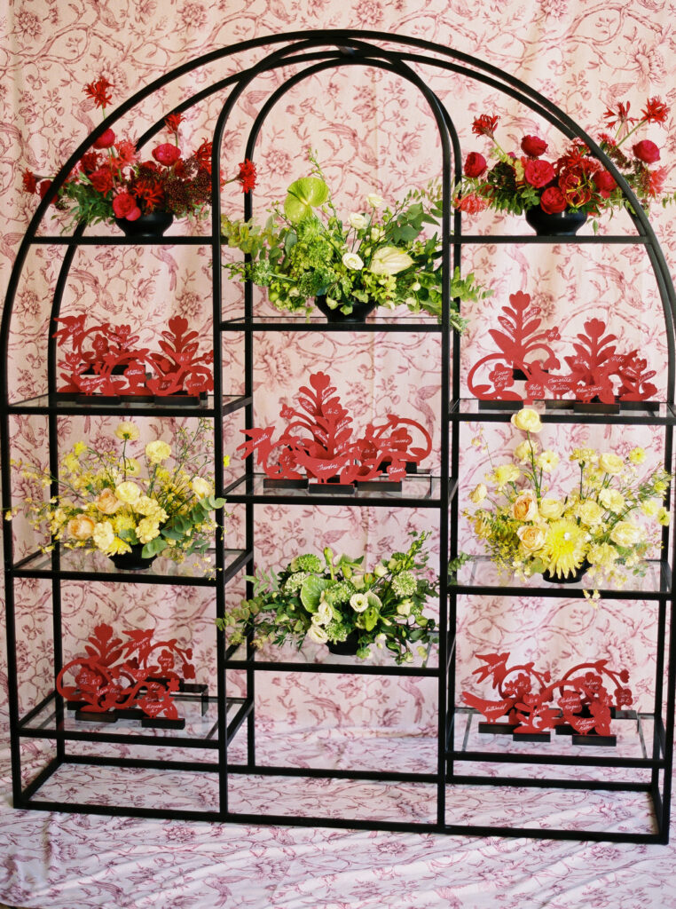 Full view of modern black shelving filled with red folk-style escort cards and colorful floral arrangements, set against a pink toile backdrop