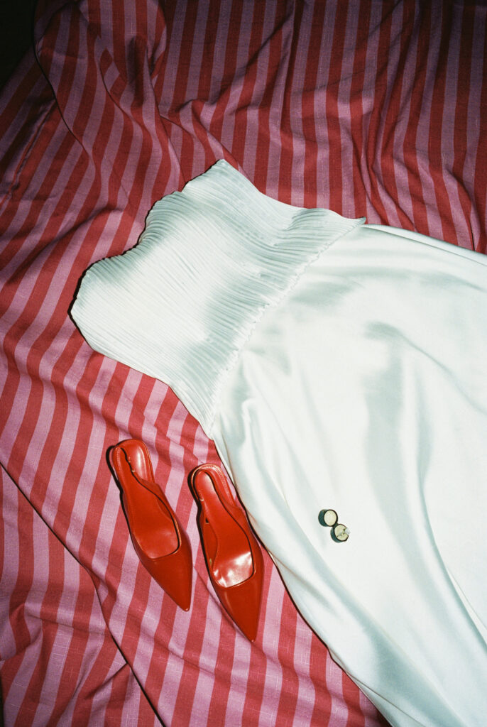 Flat lay of a white bridal gown, red Prada heels, and rings on a red striped fabric background.