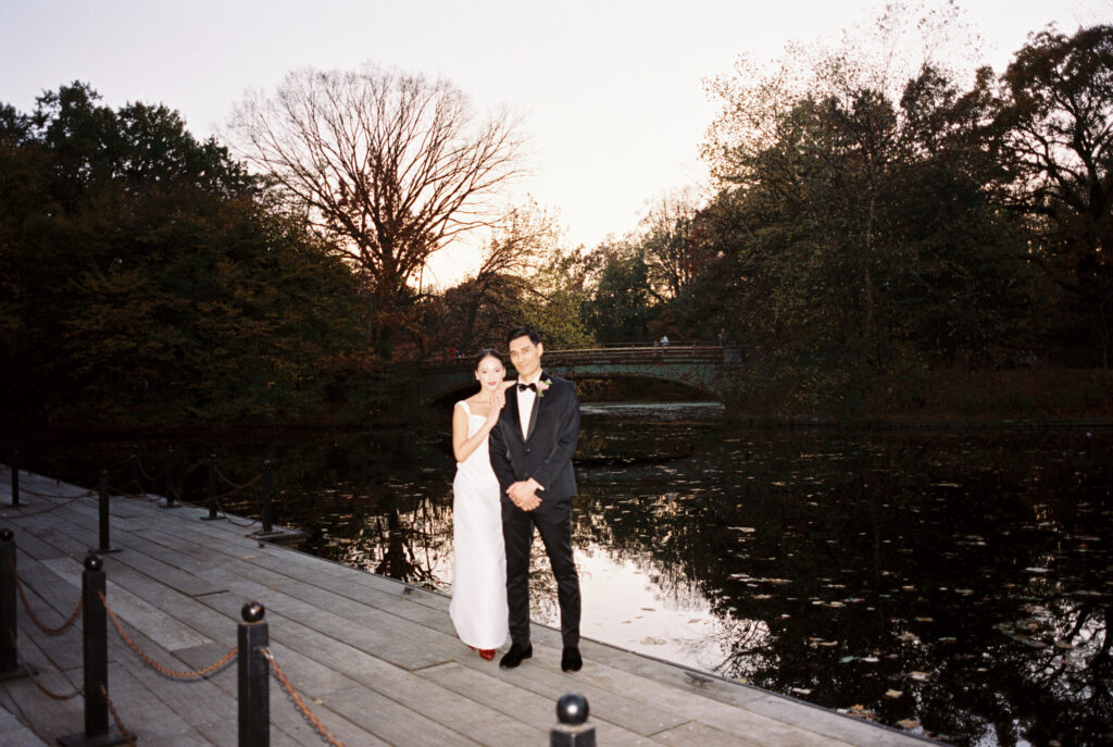 Bride and groom standing on a boardwalk by the water at sunset in Prospect Park, with a bridge in the background
